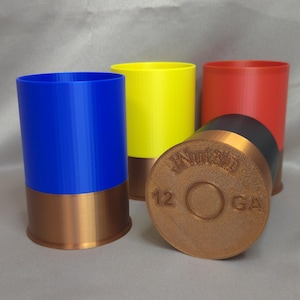 Può includere: Una collezione di contenitori cilindrici colorati che assomigliano a bossoli di fucile. I contenitori sono blu, gialli e rossi, con una base color rame. Un bossolo è sdraiato, mostrando il testo "JNuts3D" e "12 GA".