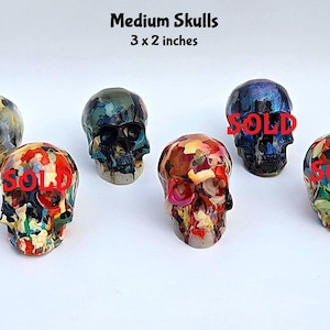 May include: Six medium skulls measuring 7.6 x 5.1 cm, featuring varied multi-coloured, marbled, and dark finishes. Two skulls are marked with the red text 'SOLD'. The skulls display detailed anatomical features including eye sockets, nasal cavities, and teeth.