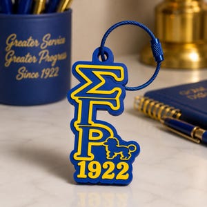 May include: A blue and yellow rubber keyring featuring the Greek letters Sigma Gamma Rho, a poodle silhouette, and the year 1922. The design is attached to a circular blue metal wire ring with a screw-top closure for securing keys.