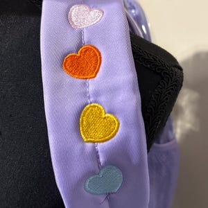 May include: A lavender backpack strap with embroidered hearts in pink, white, orange, yellow, and blue. The strap is attached to a black backpack with a rainbow-colored accent.