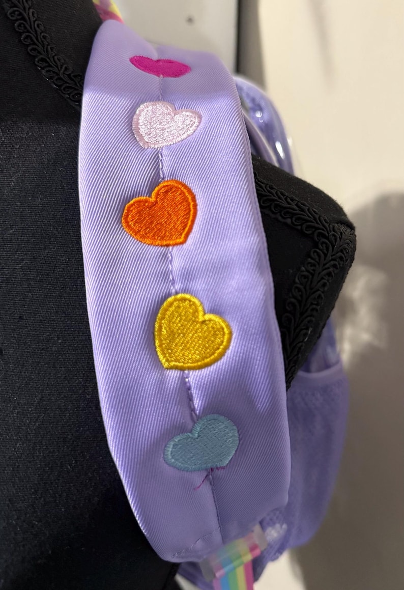 May include: A lavender backpack strap with embroidered hearts in pink, white, orange, yellow, and blue. The strap is attached to a black backpack with a rainbow-colored accent.