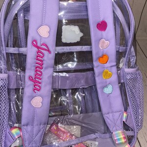 May include: A clear backpack with purple straps and accents. The straps feature embroidered hearts in various colors and the name "Jamaya" in pink script. The backpack has a rainbow-striped handle and strap details.