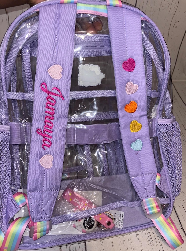May include: A clear backpack with purple straps and accents. The straps feature embroidered hearts in various colors and the name "Jamaya" in pink script. The backpack has a rainbow-striped handle and strap details.