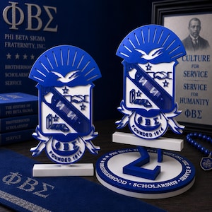 May include: Two blue and white Phi Beta Sigma fraternity crests made of layered material. Each crest features a dove, a handshake, a book, and the Greek letters ΦΒΣ with the text FOUNDED 1914. One crest stands on a circular base with the text BROTHERHOOD SCHOLARSHIP SERVICE.