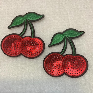 May include: Two red cherries with green stems and leaves. The cherries are made of sequins and are ideal for adding a touch of whimsy to crafts and projects.