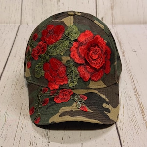 May include: Camouflage baseball cap with embroidered red roses and green leaves. The cap features a curved brim and a classic six-panel design. The floral embroidery adds a touch of elegance to the military-inspired pattern.