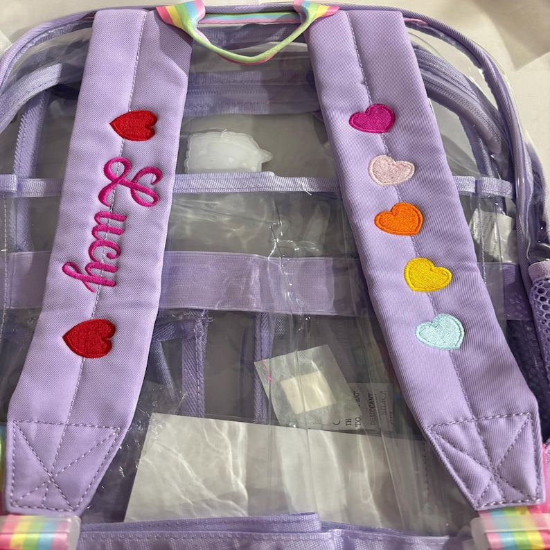 May include: A clear backpack with purple straps and accents. The left strap has the name "Lucy" embroidered in pink, with red heart accents. The right strap features colorful heart patches. The top handle has a rainbow design.