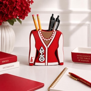 May include: A red and white desk organizer shaped like a cardigan sweater with a pearl necklace design, featuring the Greek letters Delta Sigma Theta, a small silhouette figure, and holding two yellow pencils and two black pens.