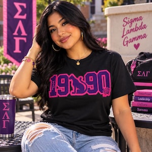Peut inclure: Un t-shirt noir arborant le texte 1990 en grandes lettres violettes et roses, avec une silhouette de panthère remplaçant le zéro et un petit motif de rose. Les lettres grecques ΣΛΓ sont imprimées à gauche du texte. Le t-shirt a un col rond.