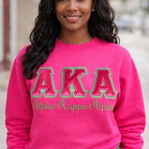 May include: A bright pink sweatshirt with the letters "AKA" in a green and pink design. Below the letters is the embroidered text "Alpha Kappa Alpha". The person in the image is wearing green pants.