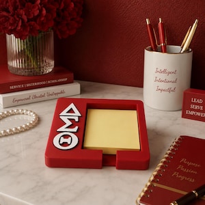 May include: A red square sticky note holder featuring the white Greek letters Delta Sigma Theta on the left side and a stack of yellow square sticky notes. The holder is surrounded by a red notebook, a white pen cup with text, and books.