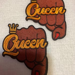 May include: Two brown embroidered patches with a raised fist and the word "Queen" in gold lettering with a gold crown above the word.