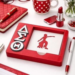 May include: A red desk organiser featuring the Greek letters ΔΣΘ and a silhouette of a person. The organiser is surrounded by a red notebook, pen, lipstick, and a polka-dot mug on a white surface.