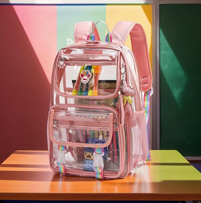 May include: A clear pink backpack with multiple compartments, including a front pocket and side mesh pocket. The backpack features rainbow-colored straps and accents, and is filled with colorful markers and a small toy. The backpack is sitting on a wooden table.