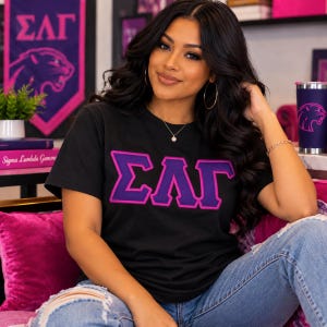 Peut inclure: Un t-shirt noir à manches courtes arborant les lettres grecques Sigma Lambda Gamma en appliqué brodé violet et rose sur la poitrine. Le vêtement présente une encolure ronde et une coupe standard, mettant en valeur les lettres en relief.