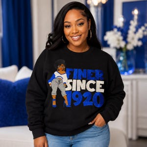 May include: A black long-sleeve crewneck sweatshirt featuring a graphic of a person wearing a white and blue top with gray pants, accompanied by the embroidered text FINER SINCE 1920 in blue and white lettering across the front of the garment.