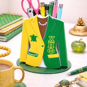 May include: A yellow and green blazer-shaped desk organizer filled with pens, pencils, and pink scissors. The organizer has a brown collar with a pearl necklace and a turtle emblem. A gold pineapple and green apple are in the background.