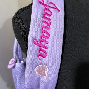 May include: Close-up of a lavender backpack strap with the name "Jamaya" embroidered in bright pink cursive. Two small, light pink heart-shaped patches are sewn onto the strap. The backpack is a light purple color.