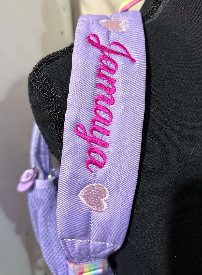 May include: Close-up of a lavender backpack strap with the name "Jamaya" embroidered in bright pink cursive. Two small, light pink heart-shaped patches are sewn onto the strap. The backpack is a light purple color.