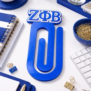 May include: A vibrant blue paperclip-shaped desk accessory with the letters ZΦB in white and blue. The image also shows a blue notebook, a keyboard, and gold-colored paperclips, all on a white surface.