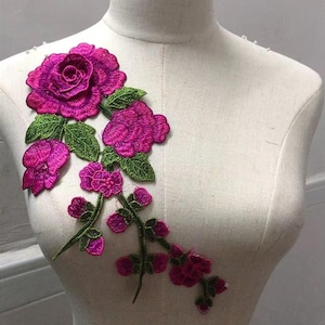 May include: A vibrant, embroidered floral applique featuring a large, magenta rose and several smaller blooms. Green leaves accent the magenta flowers. The applique is designed to be sewn onto fabric, adding a decorative touch to clothing or accessories.