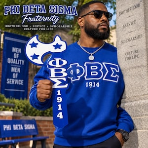 May include: A royal blue sweatshirt featuring the white Greek letters ΦΒΣ and the year 1914. The garment is held alongside a blue and white wooden axe-shaped sign displaying the same Greek letters and the year 1914, with three blue stars.