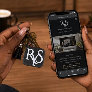 May include: A person holding a smartphone displaying the R&S Creative Studio website and a keychain with a black square tag. The tag features the white letters "R&S" and a black tassel. The phone screen shows the company logo and the text "Design. Create. Inspire."