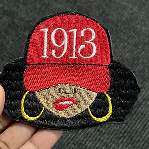 May include: Embroidered patch featuring a woman with black hair, wearing a red hat with the number 1913 embroidered on it. She is wearing large gold hoop earrings and has a red lip.