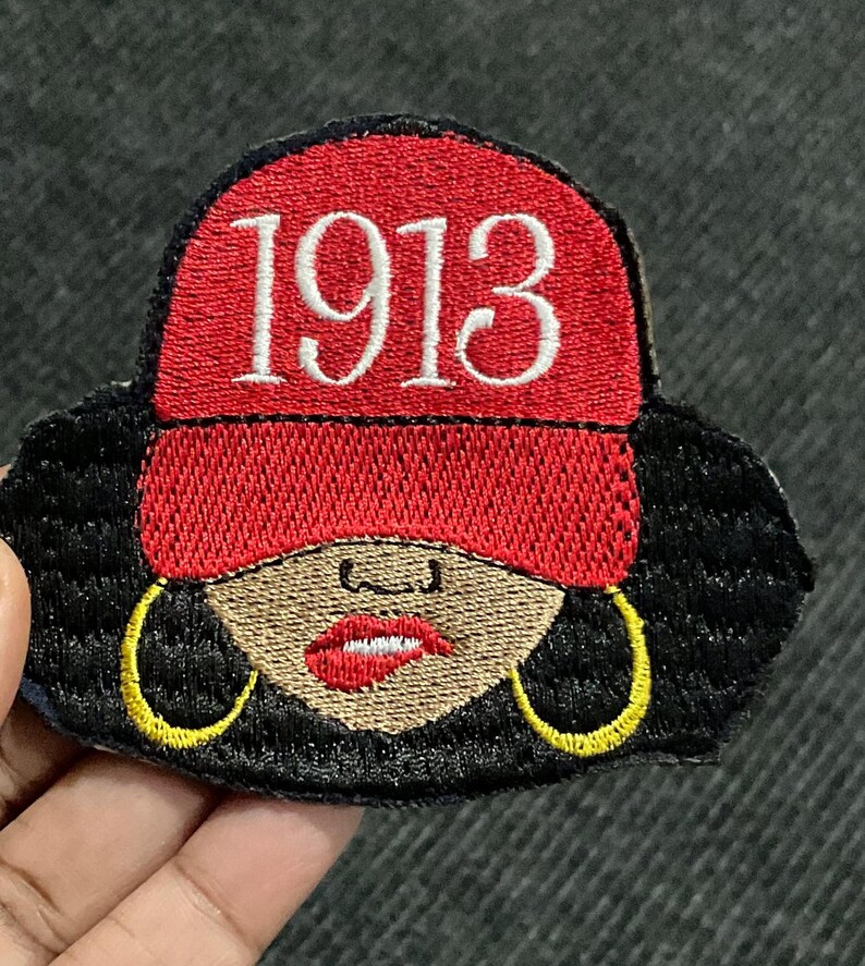 May include: Embroidered patch featuring a woman with black hair, wearing a red hat with the number 1913 embroidered on it. She is wearing large gold hoop earrings and has a red lip.