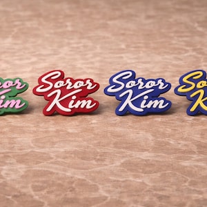 Peut inclure: Quatre épinglettes personnalisées portant le texte Soror Kim dans une police cursive. Les épinglettes sont disponibles en quatre combinaisons de couleurs : vert avec texte rose, rouge avec texte blanc, bleu avec texte blanc et bleu avec texte jaune.