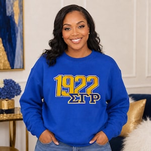 May include: A royal blue crewneck sweatshirt featuring the text 1922 in large yellow block letters with a white outline, positioned above the Greek letters ΣΓΠ in yellow with a white outline, centered on the chest of the long-sleeved garment.