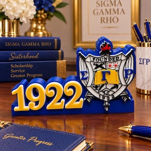 May include: A blue and yellow desk sign featuring the number 1922 and the Sigma Gamma Rho crest. The crest includes a shield with a torch, an open book, and Greek letters. The sign is displayed on a wooden surface near blue books and a pen holder.