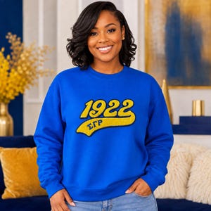 May include: A royal blue crewneck sweatshirt featuring a large, gold-colored chenille patch on the chest. The patch displays the number 1922 in a bold, curved font, with the Greek letters ΣΓΡ positioned underneath in a smaller, matching gold script.