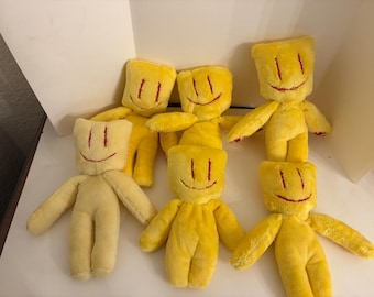 May include: A collection of six yellow plush dolls with square-shaped heads. Each doll features embroidered red vertical lines for eyes and a curved red line for a mouth. The dolls are made of soft fabric and have simple, limb-like appendages.