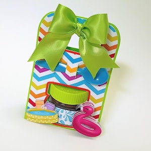 Puede incluir: Una colorida bolsa de regalo con un lazo verde, una cuchara azul y un tarro con tapa verde. La bolsa tiene un patrón de chevron en varios colores y un flotador de flamenco rosa adjunto.