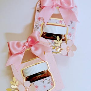 Peut inclure: Deux boîtes à dragées roses avec des motifs floraux et des accents dorés. Chaque boîte a une fenêtre transparente pour afficher un petit pot avec un couvercle brun. Les boîtes sont attachées avec des nœuds de ruban de satin rose et décorées d'une fleur rose et de feuilles dorées.