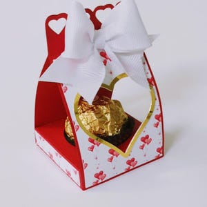 May include: A red and white gift box with a heart-shaped window, a gold-wrapped chocolate, and a large white bow. The box is decorated with red hearts and gold accents, perfect for a special occasion.