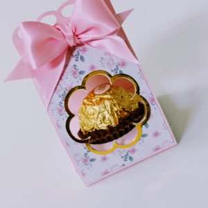 Puede incluir: Una caja de regalo rosa con un patrón floral y un recorte dorado en forma de flor. Un caramelo de chocolate es visible a través del recorte.