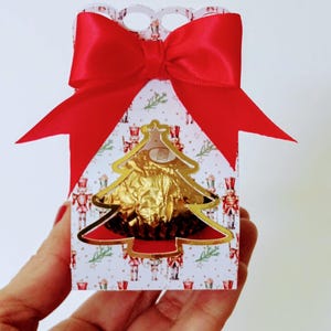 Puede incluir: Una caja de regalo de cartón blanco con forma de árbol de Navidad con un lazo rojo y un chocolate envuelto en papel dorado en su interior. La caja tiene un patrón de cascanueces.