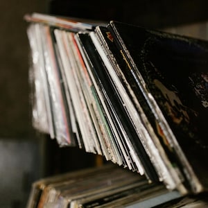 Puede incluir: Primer plano de una pila de discos de vinilo, con el disco superior visible y mostrando una imagen en blanco y negro. Los discos están dispuestos en una pila vertical, con el disco superior ligeramente desplazado de los demás. Los discos están todos en buen estado y parecen estar bien cuidados.