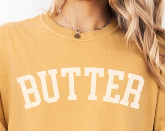 Op de afbeelding: Een mosterdgeel t-shirt met korte mouwen en het woord BUTTER in grote witte blokletters op de borst. Het shirt heeft een ronde hals en wordt gedragen met een lichtblauw spijkerjasje over de schouders gedrapeerd.