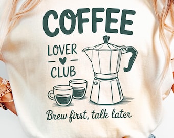 May include: Cream-colored t-shirt with a coffee-themed graphic. The design includes the word "COFFEE" above "LOVER CLUB" and an illustration of a coffee maker and two cups. The phrase "Brew first, talk later" is printed below the graphic.