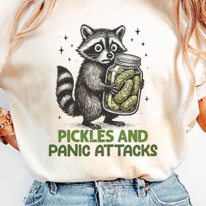 May include: Cream-colored t-shirt featuring a cartoon raccoon clutching a jar of pickles. The raccoon has a worried expression. The text "PICKLES AND PANIC ATTACKS" is printed below the raccoon in green.