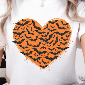 May include: A white short-sleeved t-shirt featuring a large orange heart graphic on the chest. The heart is filled with a pattern of numerous small black bats in various flight positions, creating a dense, textured appearance across the center.