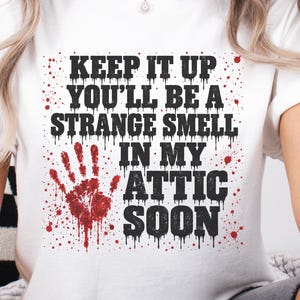 May include: A white short-sleeved t-shirt featuring black text that reads KEEP IT UP YOU'LL BE A STRANGE SMELL IN MY ATTIC SOON. The design includes a red handprint graphic and red paint splatter accents across the front of the garment.
