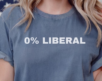 May include: A blue short-sleeved crew neck t-shirt featuring the white text 0% LIBERAL printed across the chest. The garment has a relaxed fit with rolled sleeves and is worn over blue denim jeans with a visible metal button and rivets.