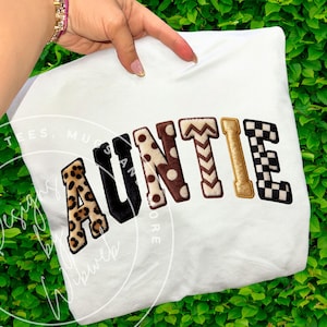 Puede incluir: Sudadera blanca con la palabra "AUNTIE" en letras coloridas y texturizadas. Las letras presentan estampado de leopardo, negro, lunares, galones, dorado y un patrón de cuadros. La sudadera se muestra sobre un fondo de follaje verde.