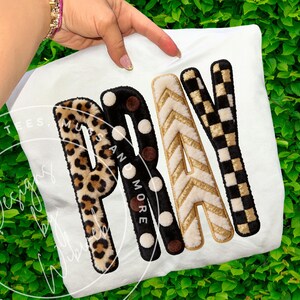 May include: White t-shirt with the word "PRAY" in bold, textured letters. The letter "P" has a leopard print, "R" has black polka dots, "A" has gold chevron, and "Y" has a black and white checkerboard pattern. The words "Mugs and More" are also visible.