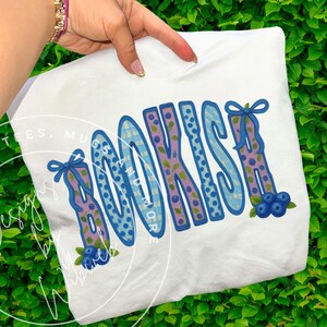 May include: White t-shirt with the word "BOOKISH" in blue and purple letters. The letters have a plaid and polka dot pattern with blueberry and leaf accents. A blue bow is on the top of the letters. The shirt is held by a person.
