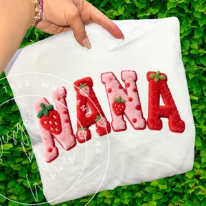 Può includere: T-shirt bianca con la scritta "NANA" in lettere rosse e rosa, a tema fragola, lettere soffici. Le lettere sono decorate con disegni di fragole. La maglietta è tenuta in mano su uno sfondo verde.